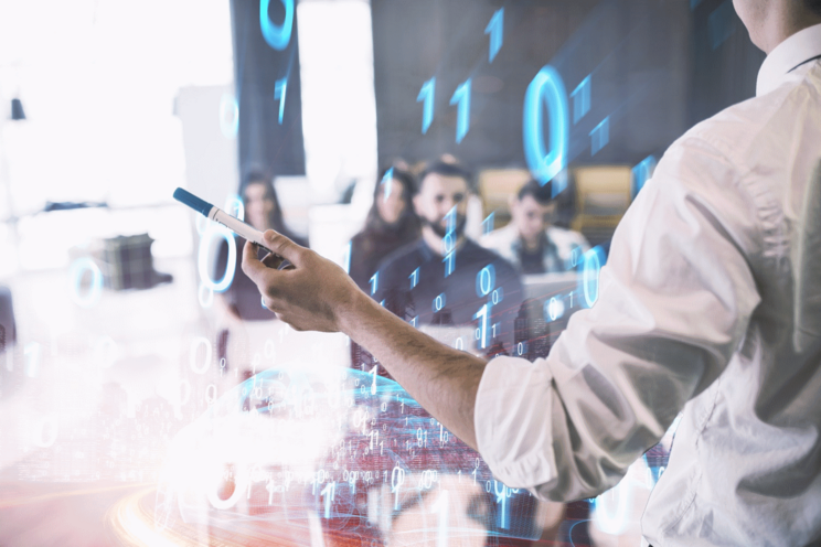 Unterrichtsszene: Dozent spricht vor Kursteilnehmenden und hält einen Stift in der Hand.  Zudem sind die Ziffern O und 1 über das Bild gelegt.