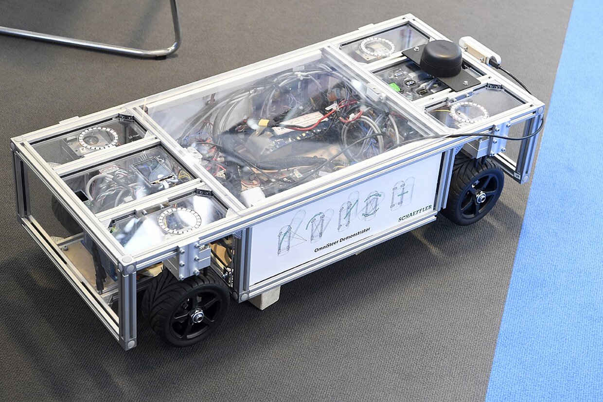 Ein viereckiges weißes Fahrzeug auf grauem Boden, rechts ragt ein blaues Dreieck ins Bild.