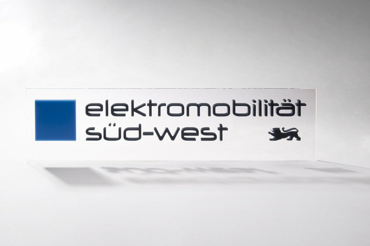 Logo Cluster Elektromobilität Süd-West Das Logo des Clusters Elektromobilität Süd-West auf einer rechteckigen Plexiglasscheibe