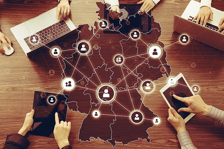 ünf Personen sitzen um einen Tisch, jeweils mit unterschiedlichen elektronischen Geräten vor sich, wie Laptops oder Tablets. In der Mitte des Tisches ist die Umrisskarte von Deutschland zu sehen. Linien verbinden die Geräte mit der Karte. Zwischen den Linien sind zahlreiche Icons von stilisierten Personen abgebildet, die Kontakte oder Netzwerke symbolisieren.