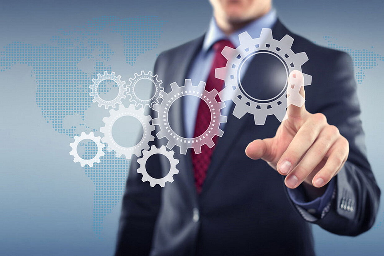 Digitalisierung Vernetzung Oberkörper einer Person im Anzug steht hinter einem Weltkartenhologramm, die Hand deutet auf eine Reihe von Zahnrädern im Vordergrund