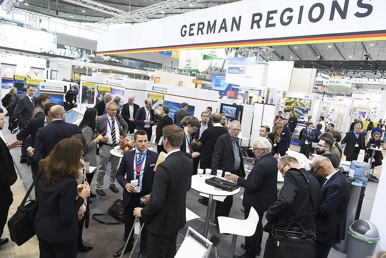 Bild auf einer Messe Mehrere Messestände aus leichter Vogelperspektive. Viele Menschen in Anzügen bewegen sich zwischen den Ständen. Über der Szene hängt ein Schriftzug mit der Aufschrift ‚German Regions‘.